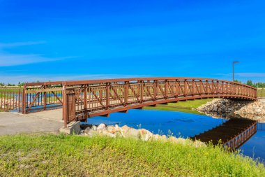 Bir köprü, her iki tarafında yemyeşil çimenli bir alanı olan bir nehir boyunca uzanır. Köprü ahşaptan yapılmıştır ve kayalarla çevrilidir. Gökyüzü berrak ve mavidir. Huzurlu ve huzurlu bir atmosfer yaratır.