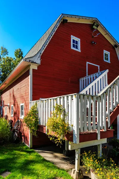 Beyaz verandalı ve basamaklı kırmızı bir ahır. Verandada saksı bitkileri ve el arabası var. Ahır yemyeşil çimlerle çevrili.