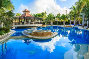 Çadırlı bir havuz ve ortasında küçük bir ada. Havuz palmiye ağaçlarıyla çevrili ve tropik bir havası var.