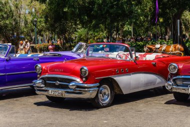 Klasik kırmızı ve beyaz bir araba bir otoparkta mor bir arabanın yanına park edilir. Kırmızı araba klasik bir model ve muhtemelen üstü açılabilir bir çatısı var.