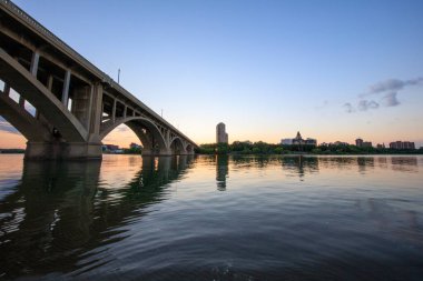 Bir köprü, arka planda bir şehir olan bir nehri kapsar. Gökyüzü turuncu ve mavi renklerin güzel bir karışımıdır.