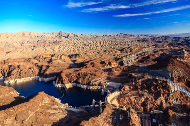 Önünde büyük bir su kütlesi ve arkasında bir köprü olan bir çöl manzarası. Gökyüzü açık ve mavidir ve uzaktaki dağlar uçsuz bucaksız ve yalnızlık hissini artırır.