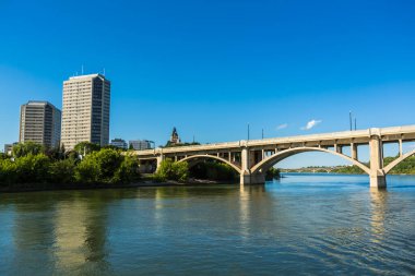 Bir köprü, arka planda bir şehir olan bir nehri kapsar. Gökyüzü açık ve mavi. Köprü büyük, beyaz bir yapıdır.