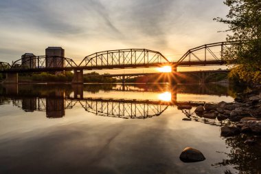 Köprü, arka planda günbatımı olan bir nehri kapsar. Köprü eski ve uzun bir geçmişi var.