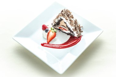 Üzerine çilek ve çikolata sosu serpiştirilmiş bir dilim pasta. Pasta ikiye bölünür ve beyaz bir tabağa konur.