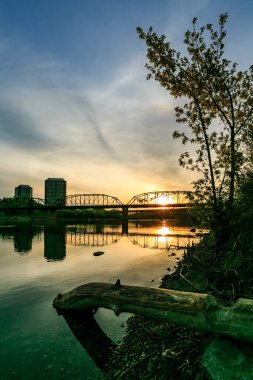 Köprü, önünde bir ağaç olan bir nehri kapsar. Güneş batıyor, sahne üzerine sıcak bir parıltı saçıyor.