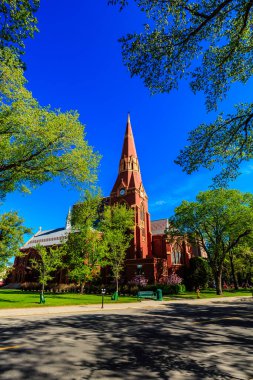 Önünde çan kulesi ve yeşil bir bank olan büyük kırmızı bir kilise. Kilise ağaçlarla çevrili ve gökyüzü mavi.