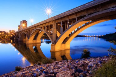 Bir köprü alacakaranlıkta bir nehri kapsar. Köprü ışıklarla aydınlanıyor, huzurlu ve huzurlu bir atmosfer yaratıyor. Suyun üzerindeki ışıkların yansıması manzaranın güzelliğini arttırır.
