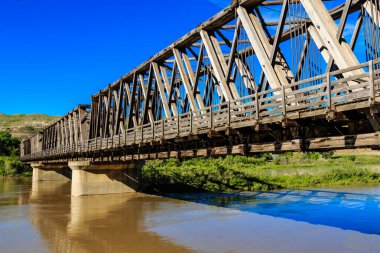Arka planda mavi gökyüzü olan bir nehir boyunca uzanan bir köprü. Köprü eski, ahşap ve su kahverengi.
