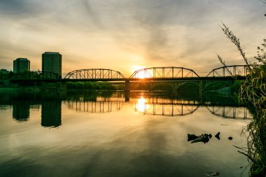 Köprü, arka planda günbatımı olan bir nehri kapsar. Köprü eski ve kırsal bir görünüme sahip.