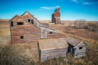 Arka planda ahır olan küçük bir ev. Ev eski ve ahır harabe halinde. Gökyüzü mavi ve çimenler kuru.
