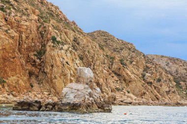 Ortasında büyük bir kaya olan kayalık bir kıyı şeridi. Kaya suyla çevrili ve gökyüzü bulutlu.