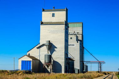 Mavi kapılı büyük bir tahıl ambarı bir tarlada oturur. Gökyüzü açık ve mavidir, ve güneş parlıyor. Silo çitlerle çevrili ve yakınlarda bir tren yolu var.