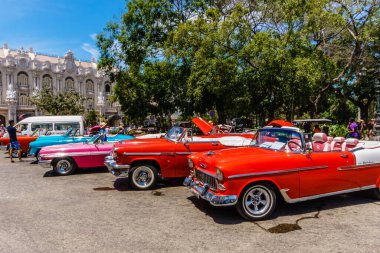 Bir binanın yanındaki otoparkta eski model arabalar var. Arabalar, pembe ve beyaz bir araba da dahil olmak üzere farklı renk ve tarzlara sahiptir. Sahne canlı ve nostaljik.