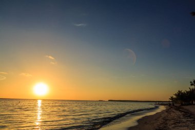 Güneş okyanusun üzerinde batıyor, plajın üzerine sıcak bir parıltı saçıyor. Gökyüzü mavi ve turuncunun bir karışımıdır. Huzurlu ve huzurlu bir atmosfer yaratır. Su sakin ve kumsal boş.
