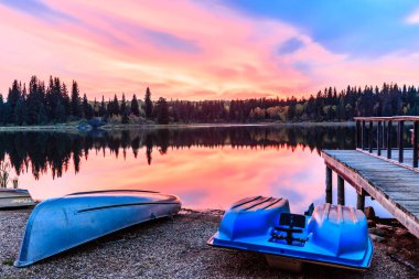 Bir gölün kıyısında mavi ve beyaz bir kano bulunur. Gökyüzü pembe ve turuncu, ve su sakin.