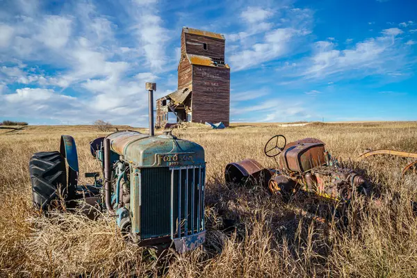 Eski bir traktör tahıl asansörünün yanındaki tarlada oturuyor. Traktör paslanmış ve silo da bakımsız durumda. Nostalji kavramı ve zaman geçişi