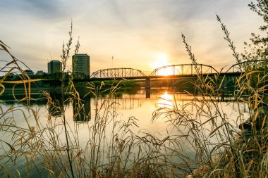 Köprü, arka planda günbatımı olan bir nehri kapsar. Köprü eski ve kırsal bir görünüme sahip. Su sakin ve güneş batıyor. Huzurlu ve sakin bir atmosfer yaratıyor.