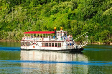 Kırmızı üst ve beyaz tabanlı büyük bir tekne bir nehirde yüzüyor. Tekne insanlarla dolu ve üzerinde 