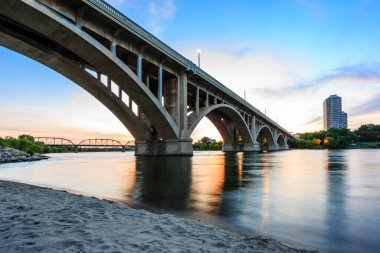Bir köprü, arka planda bir şehir olan bir nehri kapsar. Köprü ışıklarla aydınlatılıyor, huzurlu ve huzurlu bir atmosfer yaratıyor.