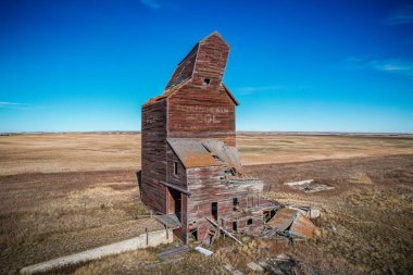 Eğik çatılı büyük, eski bir tuğla bina bir tarlada oturur. Bina terk edilmiş ve bakımsız görünüyor. Gökyüzü açık ve mavi, tarla kuru ve çorak.
