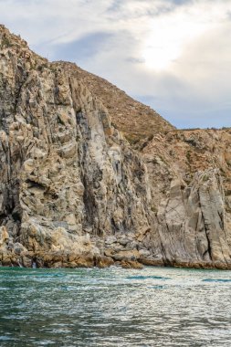 Arka planda mavi bir okyanus olan kayalık bir kıyı şeridi. Gökyüzü bulutlu ve güneş bulutların arasında parlıyor.