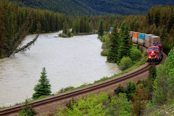 A train is traveling down a track next to a river. The train is red and is carrying a lot of cargo. The train is moving quickly and the water is choppy. The scene is peaceful and serene