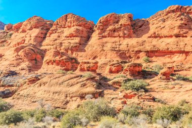 Önünde birkaç ağaç olan kayalık bir dağ sırası. Gökyüzü mavi ve güneş parlıyor.
