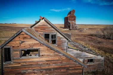 Harap olmuş eski bir ev, arka planda büyük, terk edilmiş bir tahıl ambarı olan bir tarlada oturuyor.