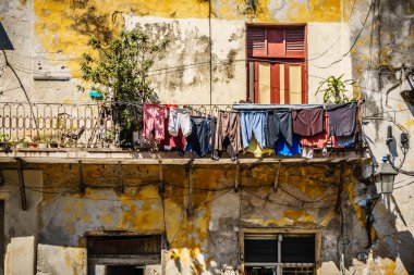 Üzerinde giysiler asılı bir balkon. Kıyafetler farklı renk ve boyutlarda. Balkon, dış görünüşü sarı ve kahverengi olan bir binanın üzerinde yer alıyor.