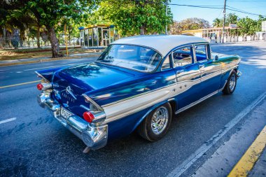 Mavi ve beyaz bir araba caddede ilerliyor. Araba eski ve klasik bir tasarımı var. Manzara nostaljik ve maziyi anımsatıyor.