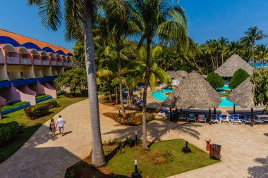 Otelin önündeki patikada yürüyen bir çift. Otelin havuzu ve palmiye ağaçları var.