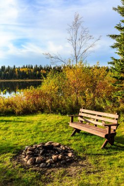 Bir gölün önünde yakındaki şömine ocağı olan ahşap bir bank oturuyor. Sahne sakin ve sakin, su ağaçları ve gökyüzünü yansıtıyor. Bankta kimse yok ama ziyaretçileri oturmaya davet ediyor.
