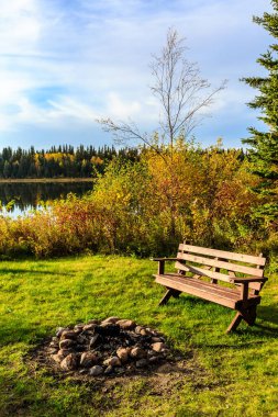Bir gölün önünde yakındaki şömine ocağı olan ahşap bir bank oturuyor. Sahne sakin ve sakin, su ağaçları ve gökyüzünü yansıtıyor. Bankta kimse yok ama ziyaretçileri oturmaya davet ediyor.