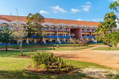 Kırmızı çatılı ve mavi çizgili büyük bir bina. Bina yemyeşil bir çim ve küçük bir bahçe ile çevrilidir. Bina, ortasında büyük bir avlusu olan bir otel ya da tatil köyü gibi görünüyor.