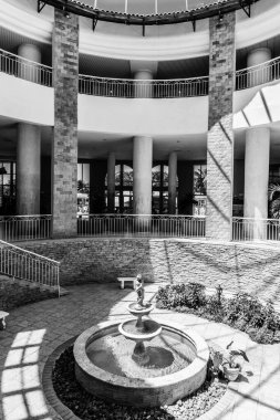 Ortasında fıskiye olan büyük bir binanın siyah beyaz fotoğrafı. Çeşme bitkiler ve bir bank ile çevrilidir. Binanın modern ve zarif bir tasarımı var.