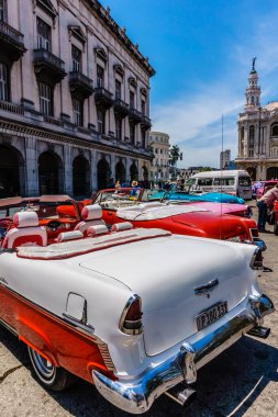 Binanın önüne bir sıra klasik araba park edilmiş. Arabaların renkleri ve stilleri farklı ve hepsi bir sırada park edilmiş. Sahne nostaljik ve klasik bir hava veriyor.