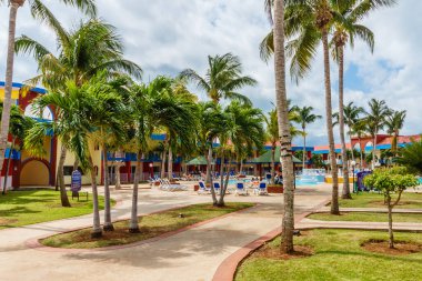 Palmiye ağaçları ve havuzu olan tropik bir tatil köyü. Tatil köyü sandalyelerde uzanan ve havuz başında dinlenen insanlarla dolu.