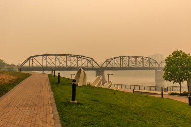 A bridge spans a river with a foggy sky in the background. The bridge is surrounded by a grassy area and a brick walkway clipart