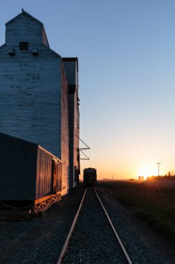 Bir tren, tahıl ambarının yanındaki raylarda ilerliyor. Güneş arka planda batıyor, sahne üzerine sıcak bir parıltı saçıyor.