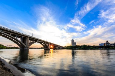 Bir köprü arka planda güzel bir günbatımı olan bir nehri kapsar. Gökyüzü açık ve güneş batıyor, suyun üzerine sıcak bir parıltı saçıyor. Köprü bir bağlantı ve ilerleme sembolüdür.