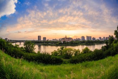 Bir şehrin ufuk çizgisi içinden akan bir nehirle uzaktan görülebilir. Gökyüzü mavi ve turuncu renklerin karışımıdır. Huzurlu ve huzurlu bir atmosfer yaratır.