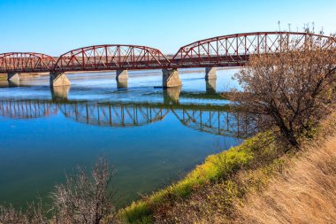 A bridge spans a river with a reflection of the bridge in the water. The bridge is red and the water is blue clipart