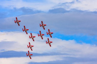 Bir grup kırmızı uçak gökyüzünde düzen içinde uçuyor. Düzen bir haç şeklinde ve uçaklar sırayla uçuyor. Gökyüzü bulutlu, ama uçaklar hala uçabiliyor.
