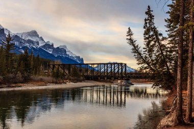 Bir köprü, arka planında güzel bir dağ sırasına sahip bir nehri kapsar. Köprü eski ve kırsal bir görünüme sahip. Su sakin ve çevresindeki manzarayı yansıtıyor.