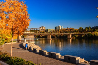 Bir köprü, arka planda bir şehir olan bir nehri kapsar. Köprü kaldırımlı ve banklı bir park ile çevrilidir. Gökyüzünü ve ağaçları yansıtan suyla sahne huzurlu ve huzurlu.