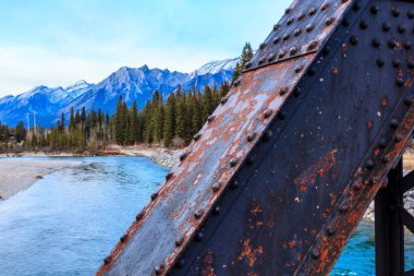 A bridge with a rusted metal frame is over a river. The bridge is old and rusted, and the water below is calm and clear. The mountains in the background add to the serene atmosphere of the scene clipart