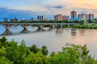 Üzerinden bir nehir akan ve üzerinde bir köprü olan bir şehir. Köprü büyüktür ve nehrin genişliğini kapsar. Şehir hareketle dolup taşıyor ve gökyüzü mavi ve turuncu karışımı.