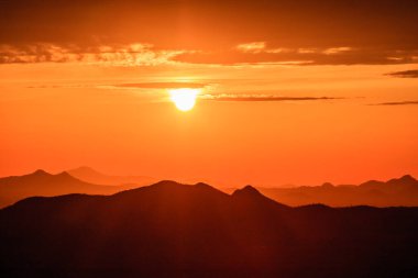 Güneş bir dağ sırasının üzerinde batıyor, manzaranın üzerine sıcak bir turuncu parıltı saçıyor. Gökyüzü bulutlarla dolu. Huzurlu ve huzurlu bir atmosfer yaratıyor.