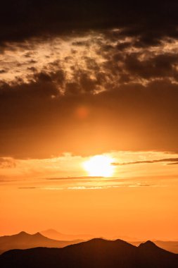 Güneş bir dağ sırasının üzerinde batıyor, manzaranın üzerine sıcak bir parıltı saçıyor. Gökyüzü bulutlarla dolu, karamsar bir atmosfer yaratıyor.
